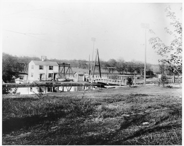 This photograph shows both the Landing Lane swing bridge (Bridge No. 26) over the Delaware and Raritan Canal in New Brunswick, as well as the bridgetender’s house and shelter.  Beyond the A-Frame bridge the steel truss bridge which carried traffic over the Raritan River to Piscataway can be seen.  The wooden A-Frame bridge was eventually replaced by the Pennsylvania Railroad during their management of the canal with the cantilevered pony plate girder (inoperative) swing bridge that remains in place to this day.